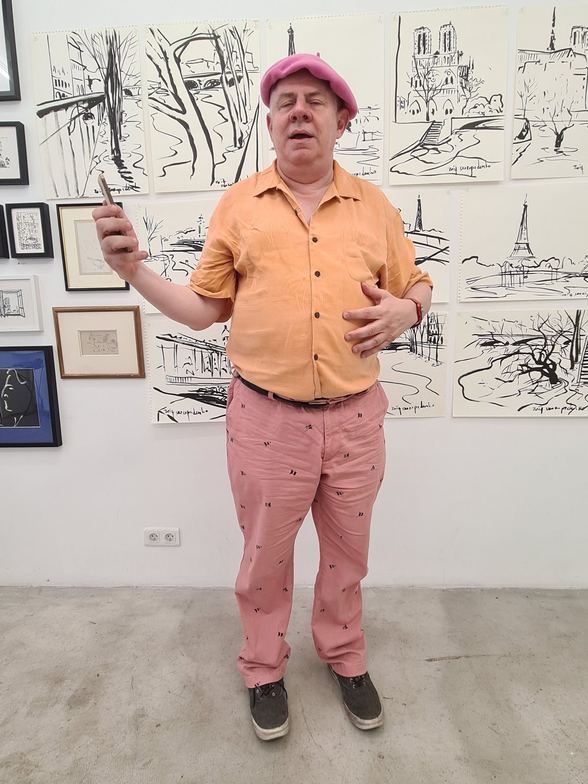 Clem Chambers in a pink beret and orange shirt gesturing in a gallery with landmark drawings on the wall