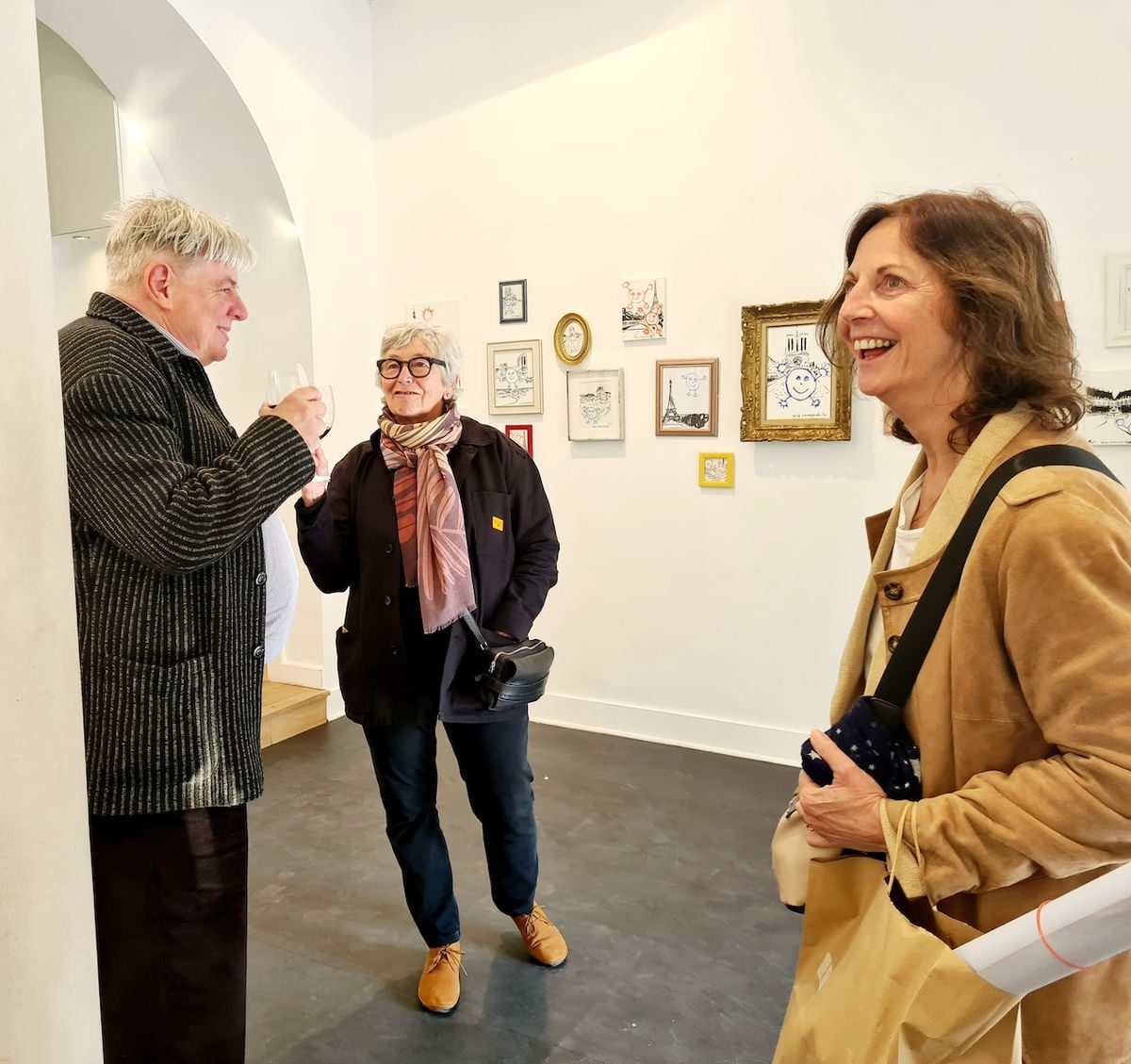 Three people laughing and talking at the Paris gallery opening with artworks on the wall behind